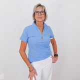 Woman wearing a light blue polo shirt and white pants against a plain background