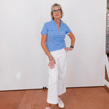 Woman wearing a blue shirt and white pants standing against a white wall.