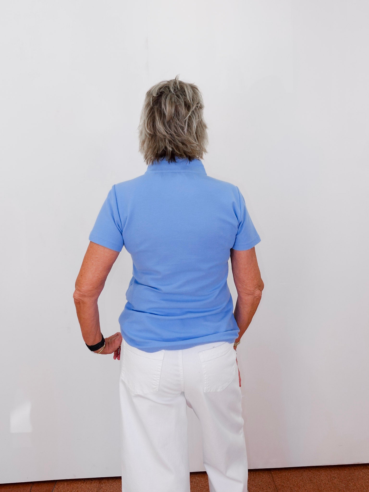 Person wearing a blue polo shirt and white pants standing against a white wall.
