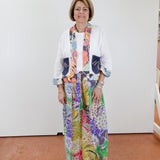 Woman wearing a colorful outfit with a patterned scarf and pants against a white wall.