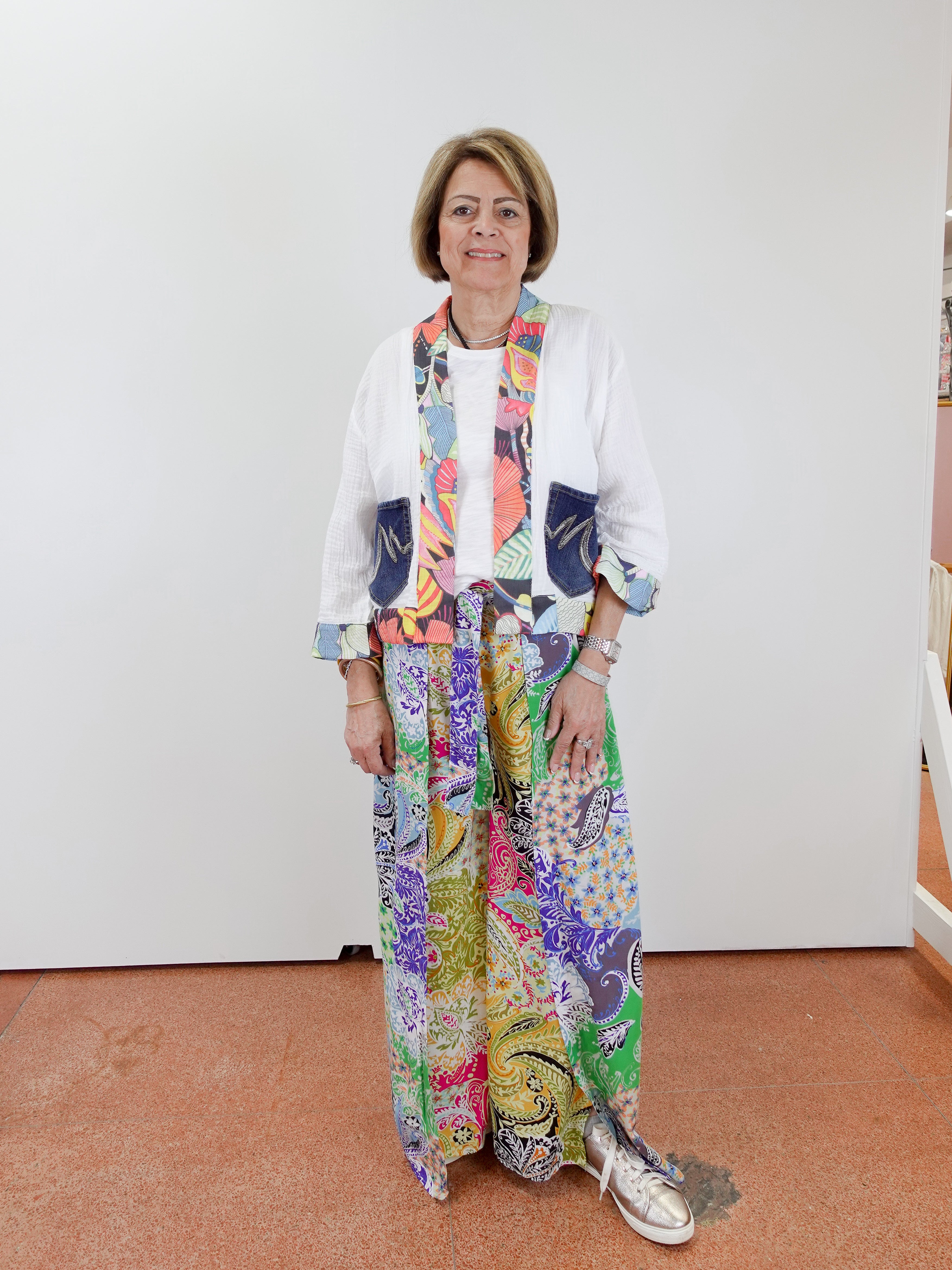 Woman wearing a colorful outfit with a patterned scarf and pants against a white wall.