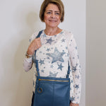 Woman wearing a star-patterned shirt and blue overalls holding a blue bag indoors.