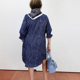 A woman viewed from behind wearing a navy blue patterned shirt dress with a white collar and a black-and-white patterned neck scarf. She is holding a light blue handbag and wearing black platform sandals, standing against a plain white wall on a terracotta tiled floor.