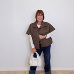 Woman wearing a brown puffer vest, white shirt, blue jeans, and brown shoes, holding a white handbag against a white wall.