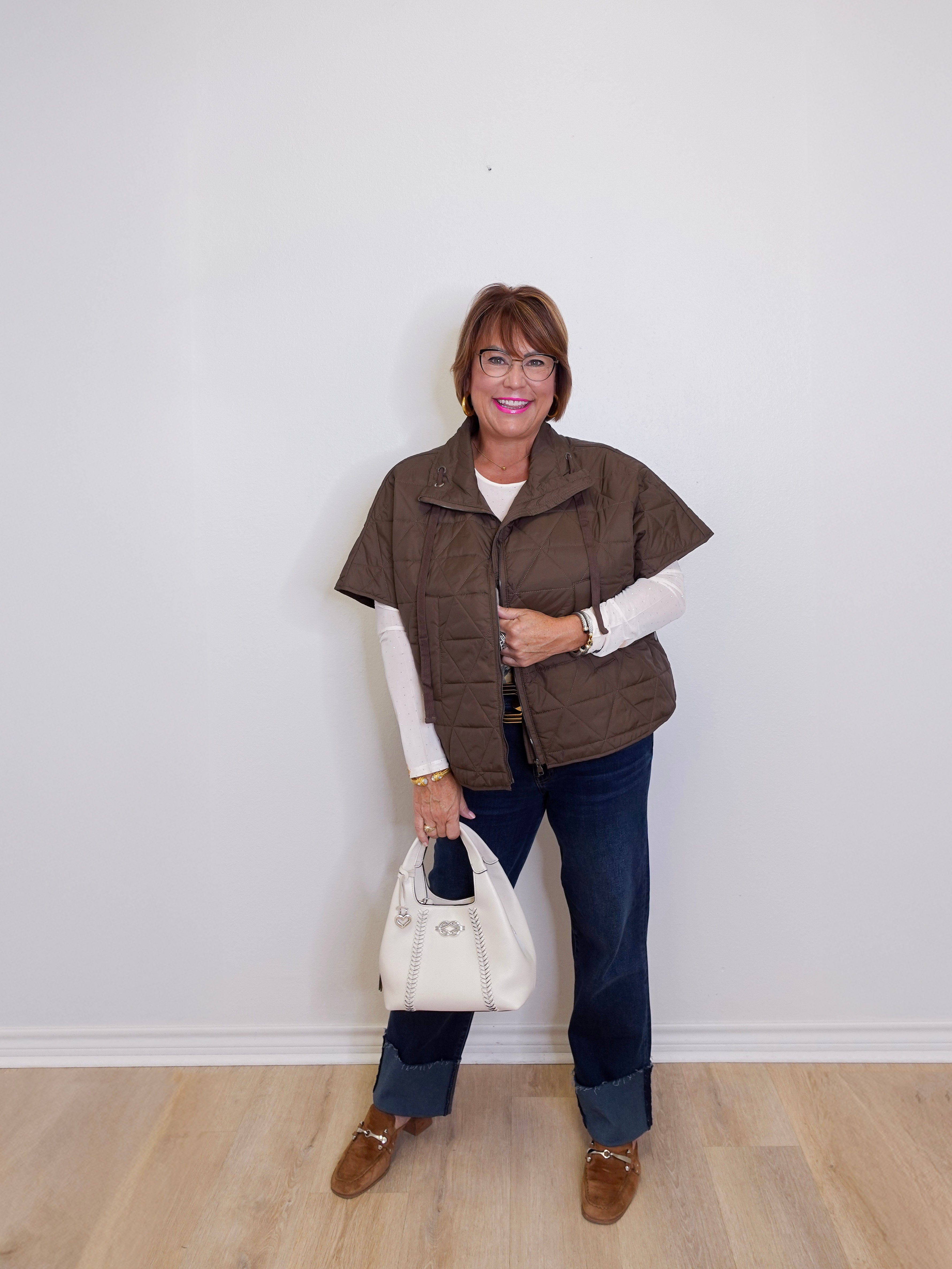 Woman wearing a brown puffer vest, white shirt, blue jeans, and brown shoes, holding a white handbag against a white wall.