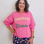 Woman wearing a pink sweatshirt with 'Champagne & Mistletoe' text and patterned shorts on a white background