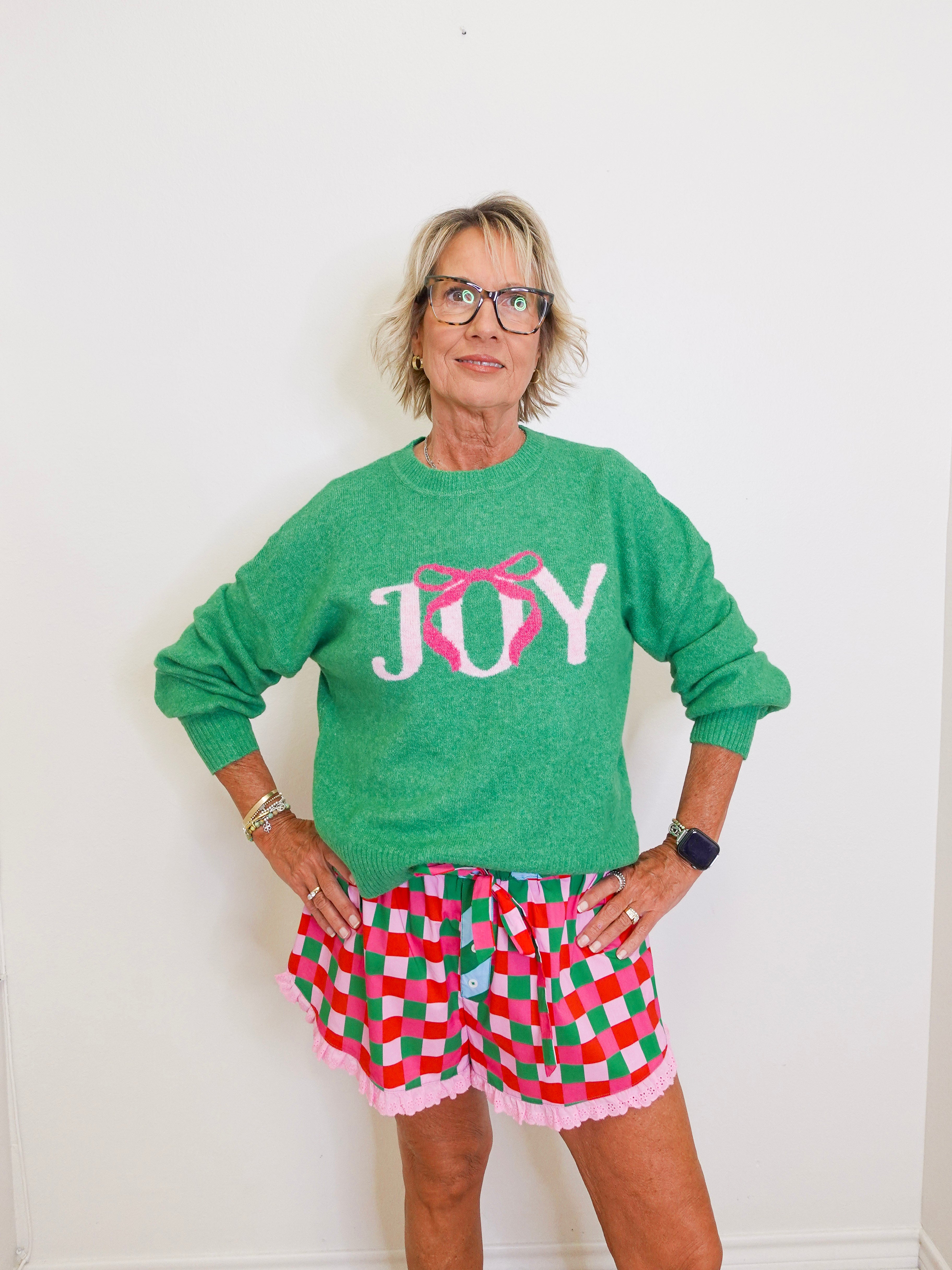 Person wearing a green 'JOY' sweater and checkered shorts against a white background