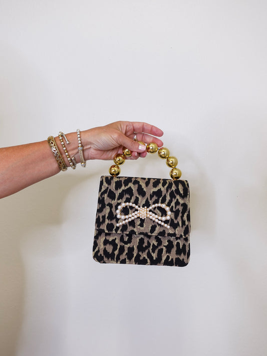 Leopard print handbag with a bow and gold handle held by a hand against a white background