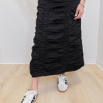 Person wearing a black ruched skirt with white sneakers on a light wooden floor.