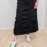 Person wearing a black ruched skirt with white sneakers on a light wooden floor.