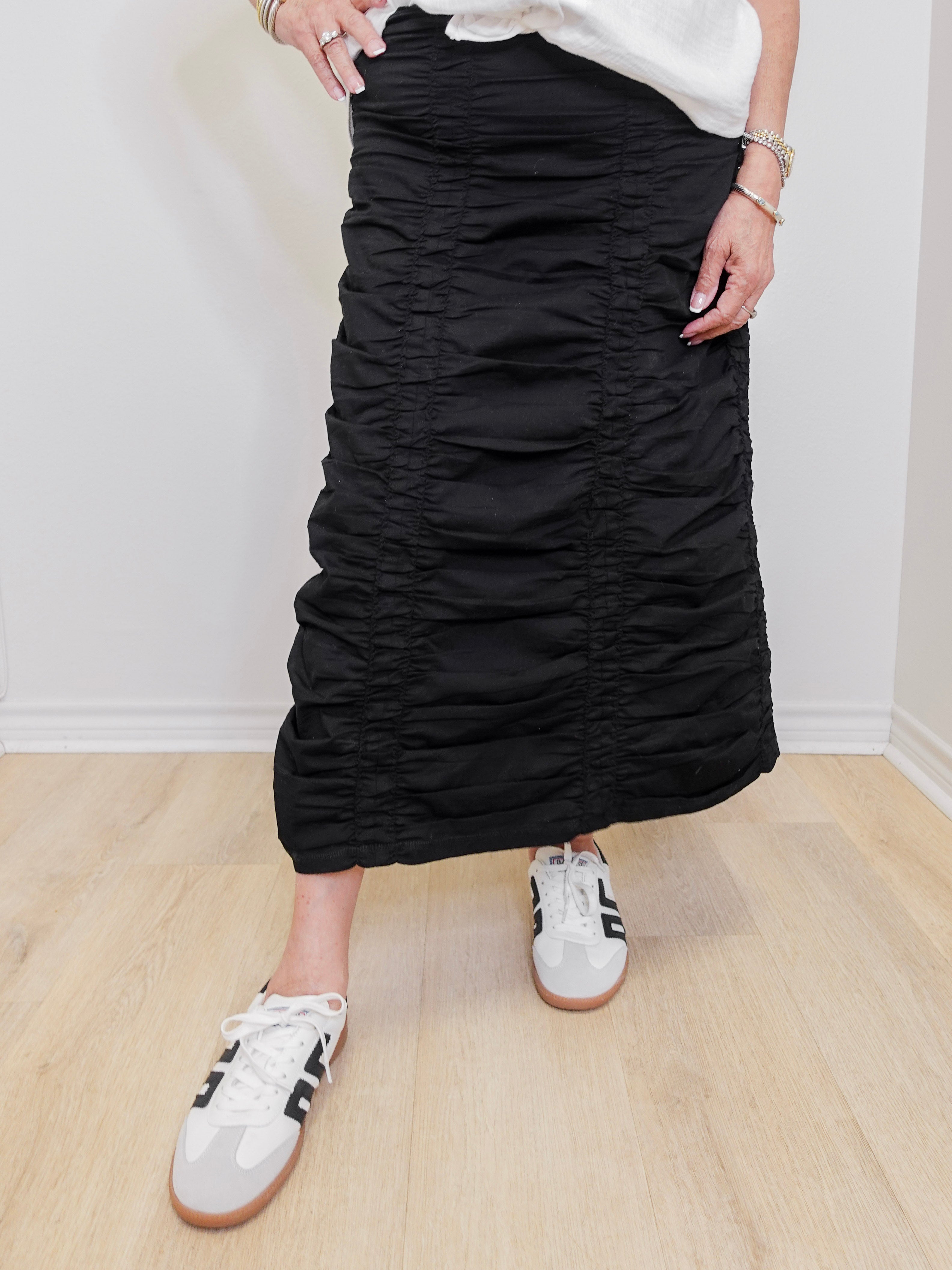 Person wearing a black ruched skirt with white sneakers on a light wooden floor.
