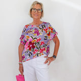 Woman wearing a colorful floral blouse and white pants, holding a pink clutch against a white background