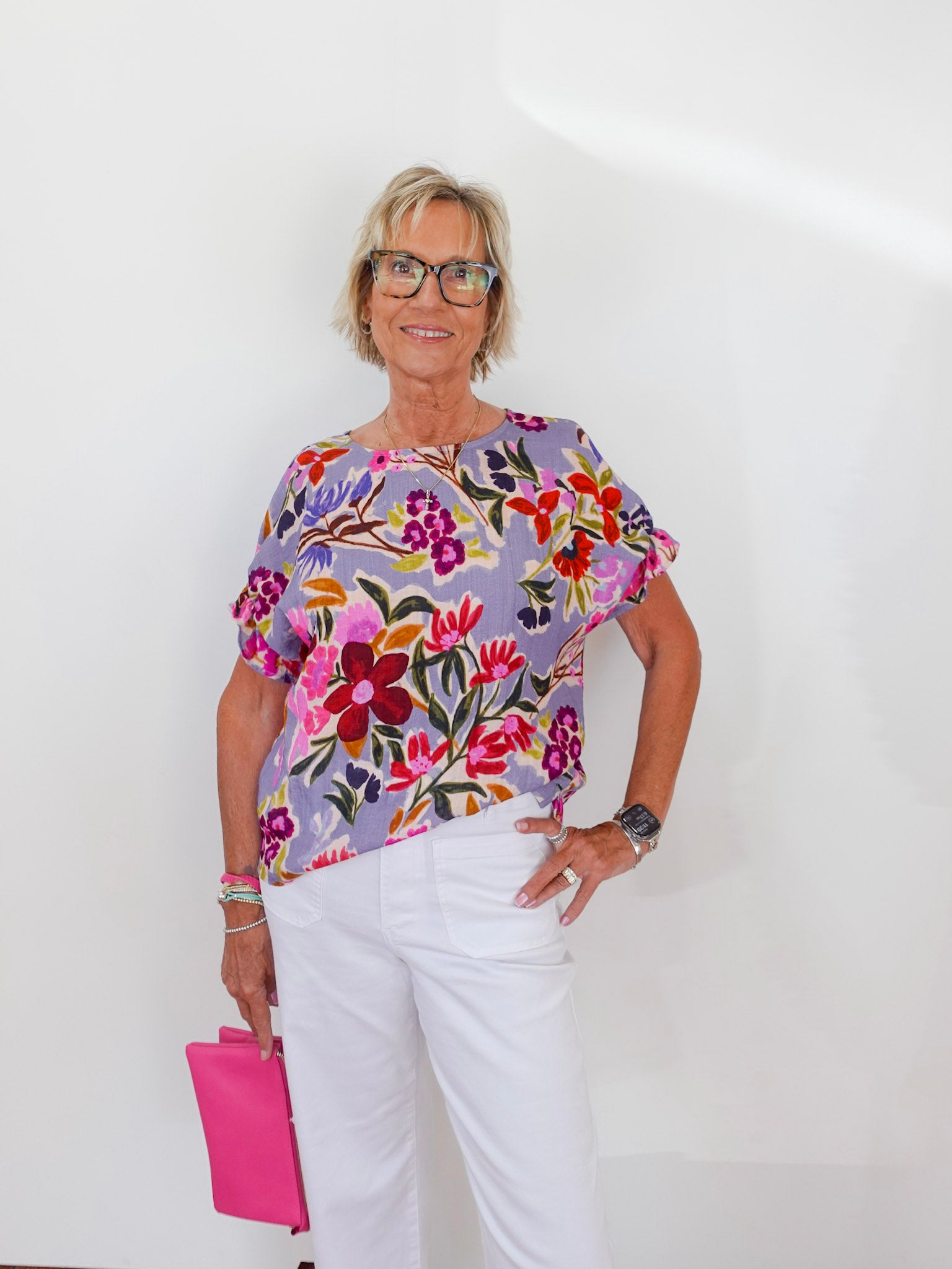 Woman wearing a colorful floral blouse and white pants, holding a pink clutch against a white background