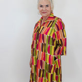 A woman modeling a short-sleeve collared shirt dress with a colorful abstract block pattern in red, orange, yellow, green, and brown against a plain white wall.