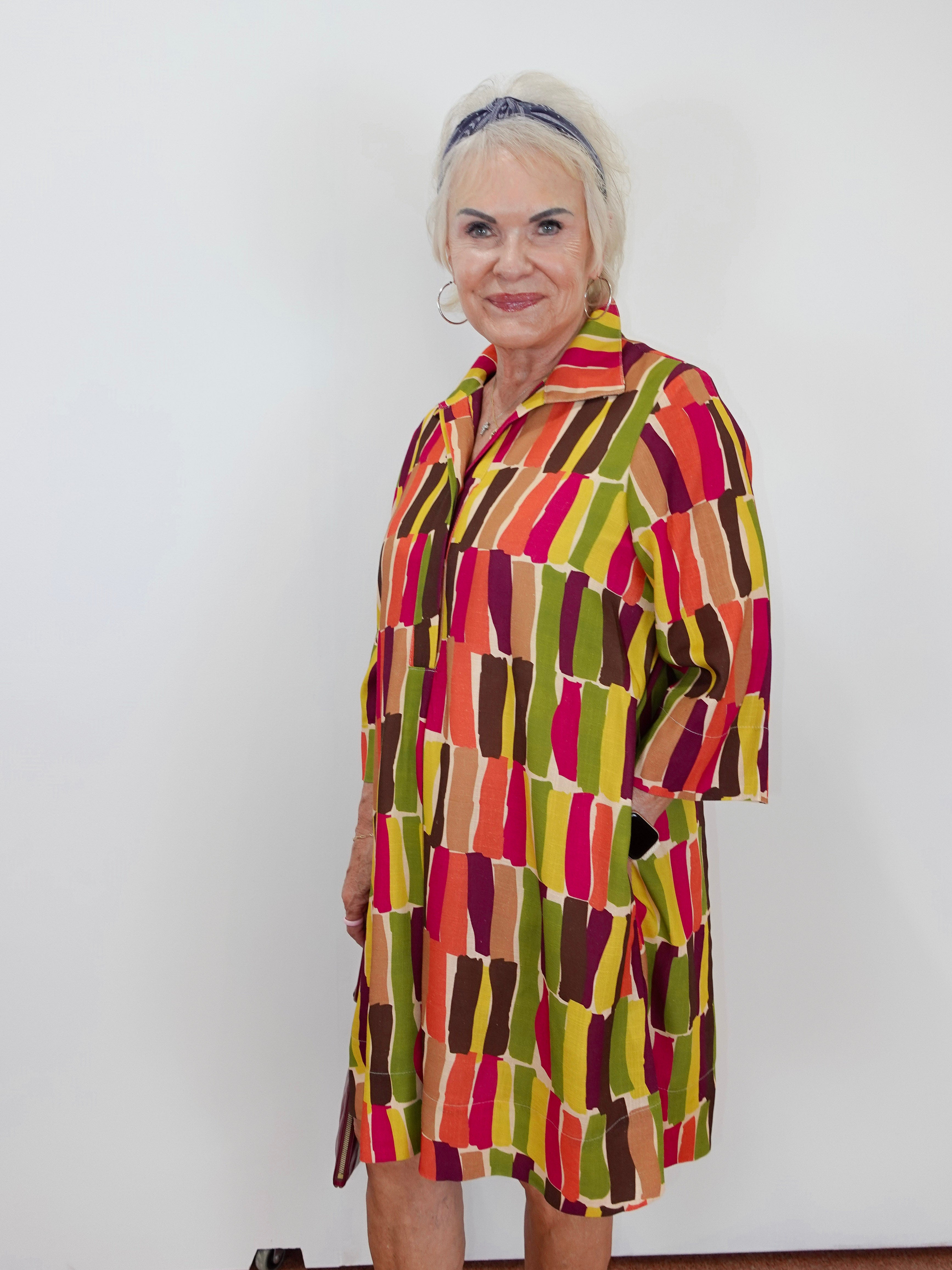 A woman modeling a short-sleeve collared shirt dress with a colorful abstract block pattern in red, orange, yellow, green, and brown against a plain white wall.