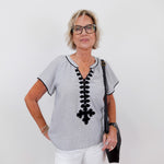 Woman wearing a gray blouse with black lace details and white pants on a plain background