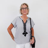 Woman wearing a gray blouse with black lace details and white pants on a plain background