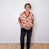 A women wearing a beige short sleeve floral print top with black pants and gold shoes against a white wall. 