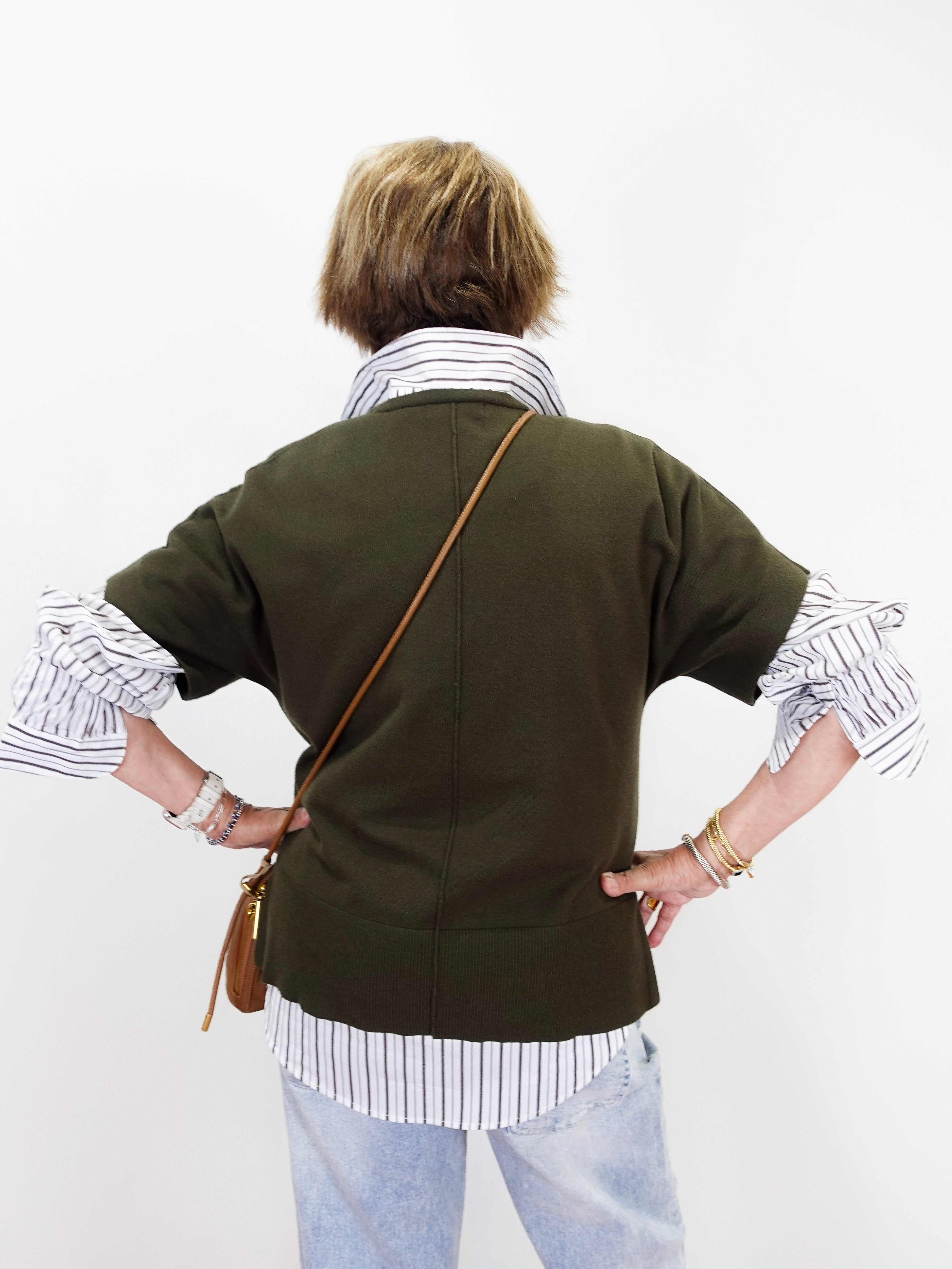 A person shown from the back wearing an olive green short-sleeve cardigan over a white and navy striped shirt, paired with light blue jeans and a brown leather crossbody bag, against a plain white background.