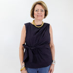 Woman wearing a black sleeveless top with a knot detail and blue pants against a white background