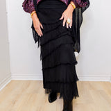 Person wearing a black ruffled skirt with a purple top and black shoes on a light wood floor.