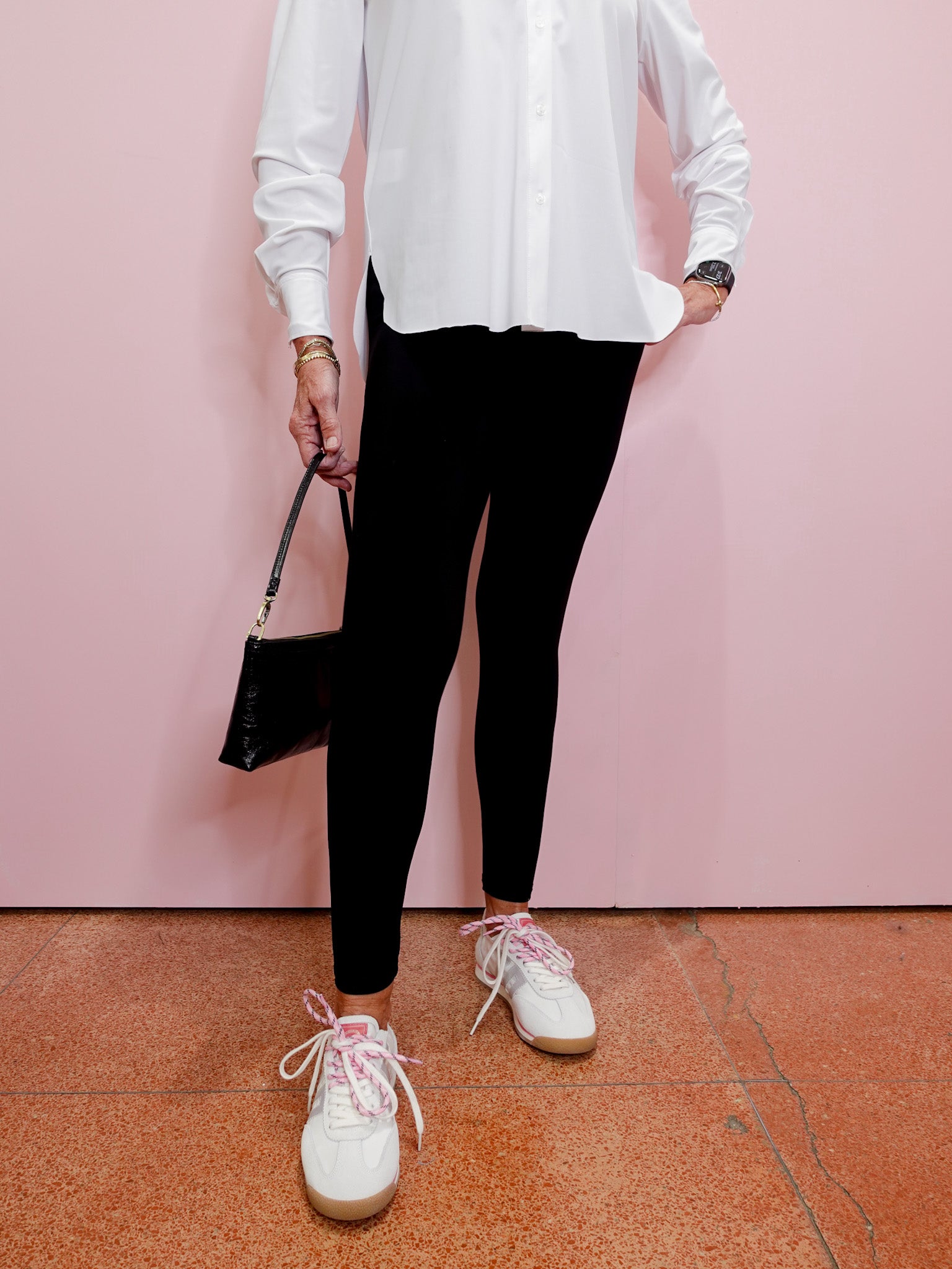Person wearing a white shirt, black leggings, and white sneakers against a pink wall.