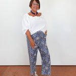 Woman wearing a white blouse and blue patterned pants against a white wall.