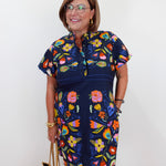 A woman wearing a navy blue short-sleeve dress with a colorful floral print, holding a beige tote bag against a plain white wall.