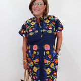 A woman wearing a navy blue short-sleeve dress with a colorful floral print, holding a beige tote bag against a plain white wall.