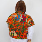 A woman viewed from behind wearing a vibrant orange short-sleeve button-up shirt with a colorful Frida Kahlo-inspired print, layered over a white long-sleeve shirt and black pants.