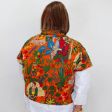 A woman viewed from behind wearing a vibrant orange short-sleeve button-up shirt with a colorful Frida Kahlo-inspired print, layered over a white long-sleeve shirt and black pants.