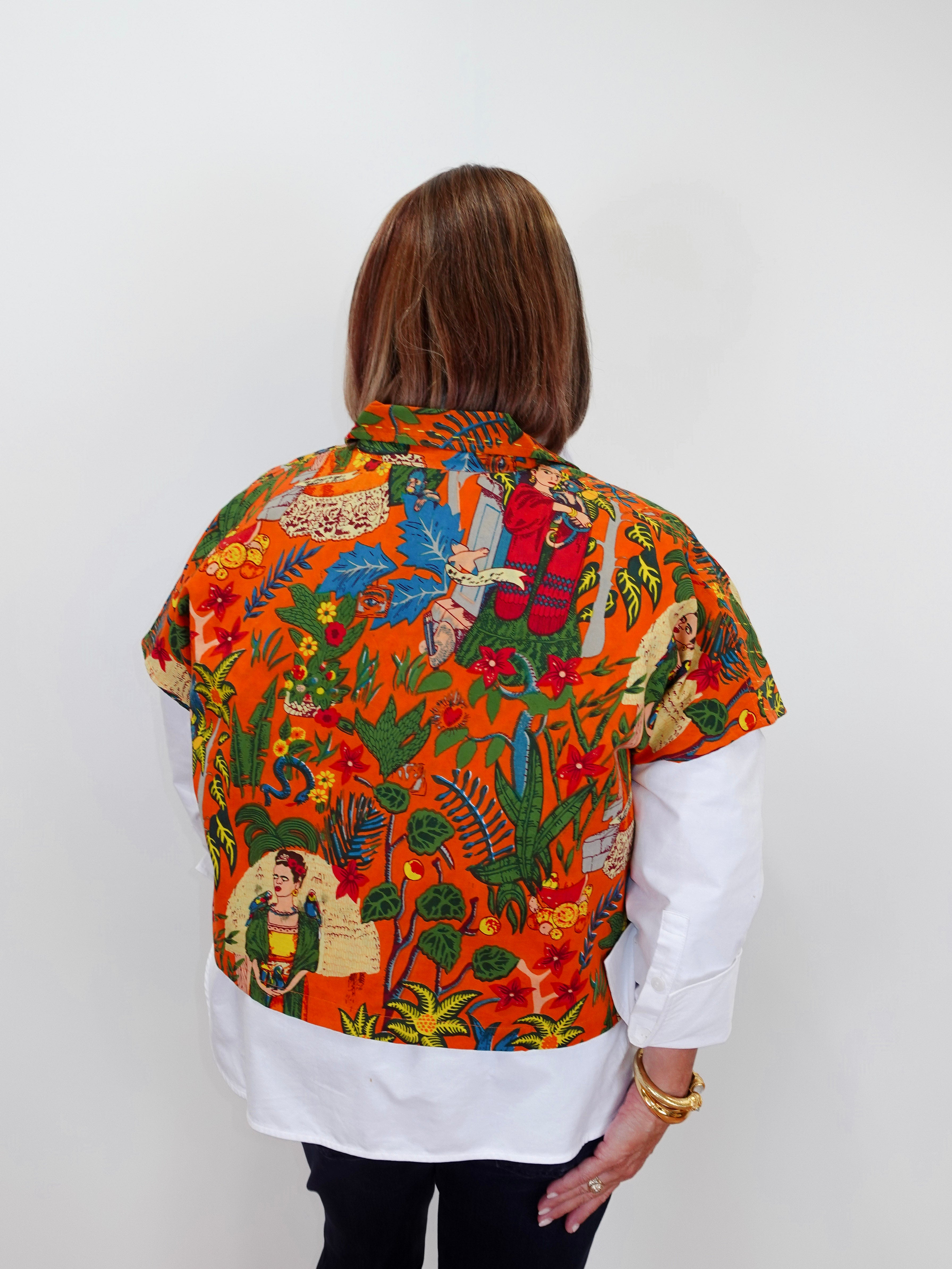 A woman viewed from behind wearing a vibrant orange short-sleeve button-up shirt with a colorful Frida Kahlo-inspired print, layered over a white long-sleeve shirt and black pants.