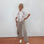 A woman stands against a white wall wearing a white short-sleeve button-up shirt with colorful cartoon graphics on the chest, beige cropped trousers, a wide gold belt, white sneakers, and carrying a small taupe handbag.