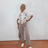A woman stands against a white wall wearing a white short-sleeve button-up shirt with colorful cartoon graphics on the chest, beige cropped trousers, a wide gold belt, white sneakers, and carrying a small taupe handbag.