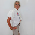 A woman with short gray hair and glasses stands against a plain white wall, wearing a white short-sleeve button-up blouse with colorful embroidered details on the chest and beige trousers.