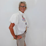 A woman with short gray hair and glasses stands against a plain white wall, wearing a white short-sleeve button-up blouse with colorful embroidered details on the chest and beige trousers.