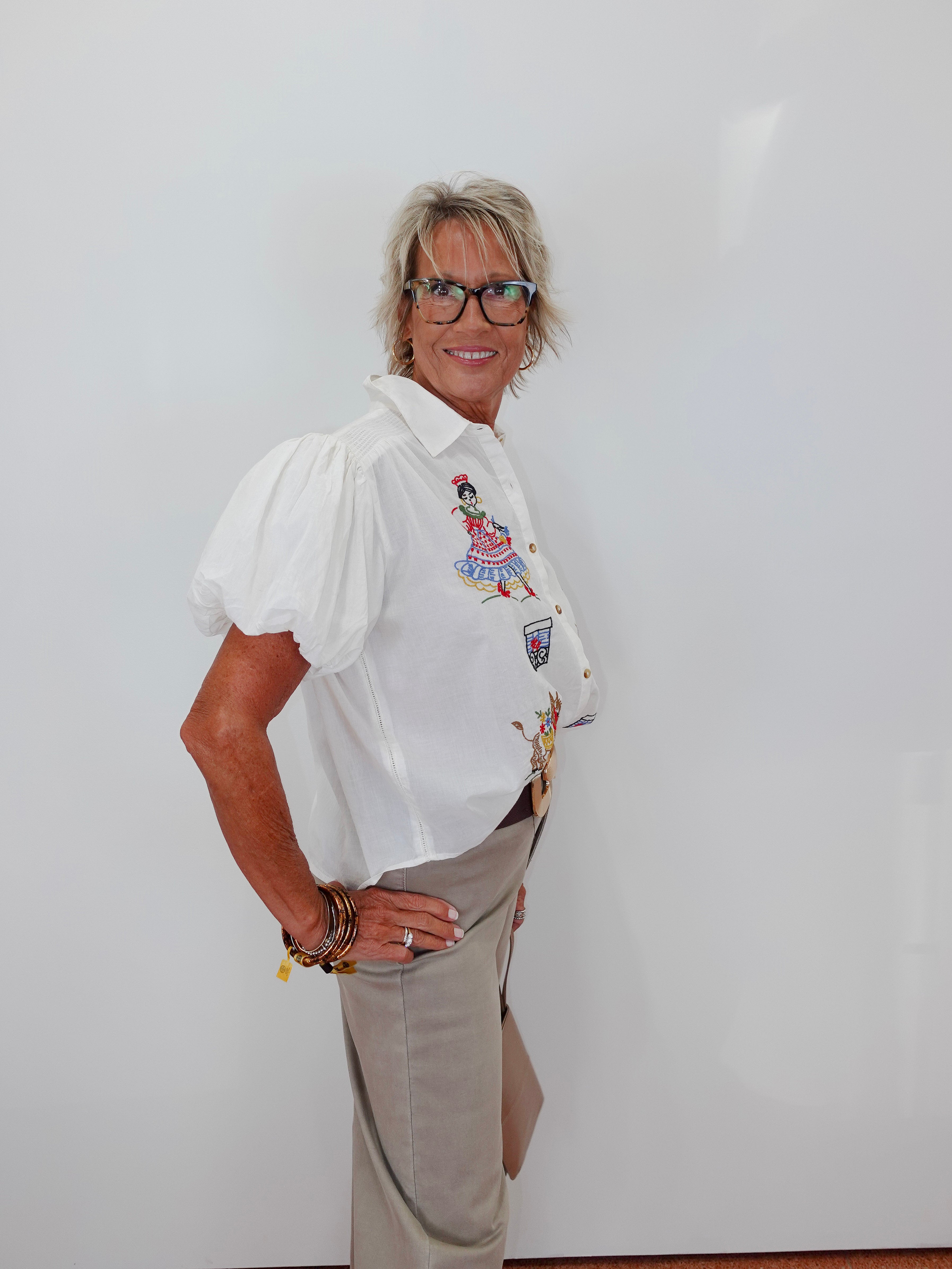 A woman with short gray hair and glasses stands against a plain white wall, wearing a white short-sleeve button-up blouse with colorful embroidered details on the chest and beige trousers.