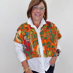 A woman wearing a short-sleeve button-up shirt with a vibrant Mexican Tehuana-style print in orange, green, and red, layered over a white collared shirt and dark pants.