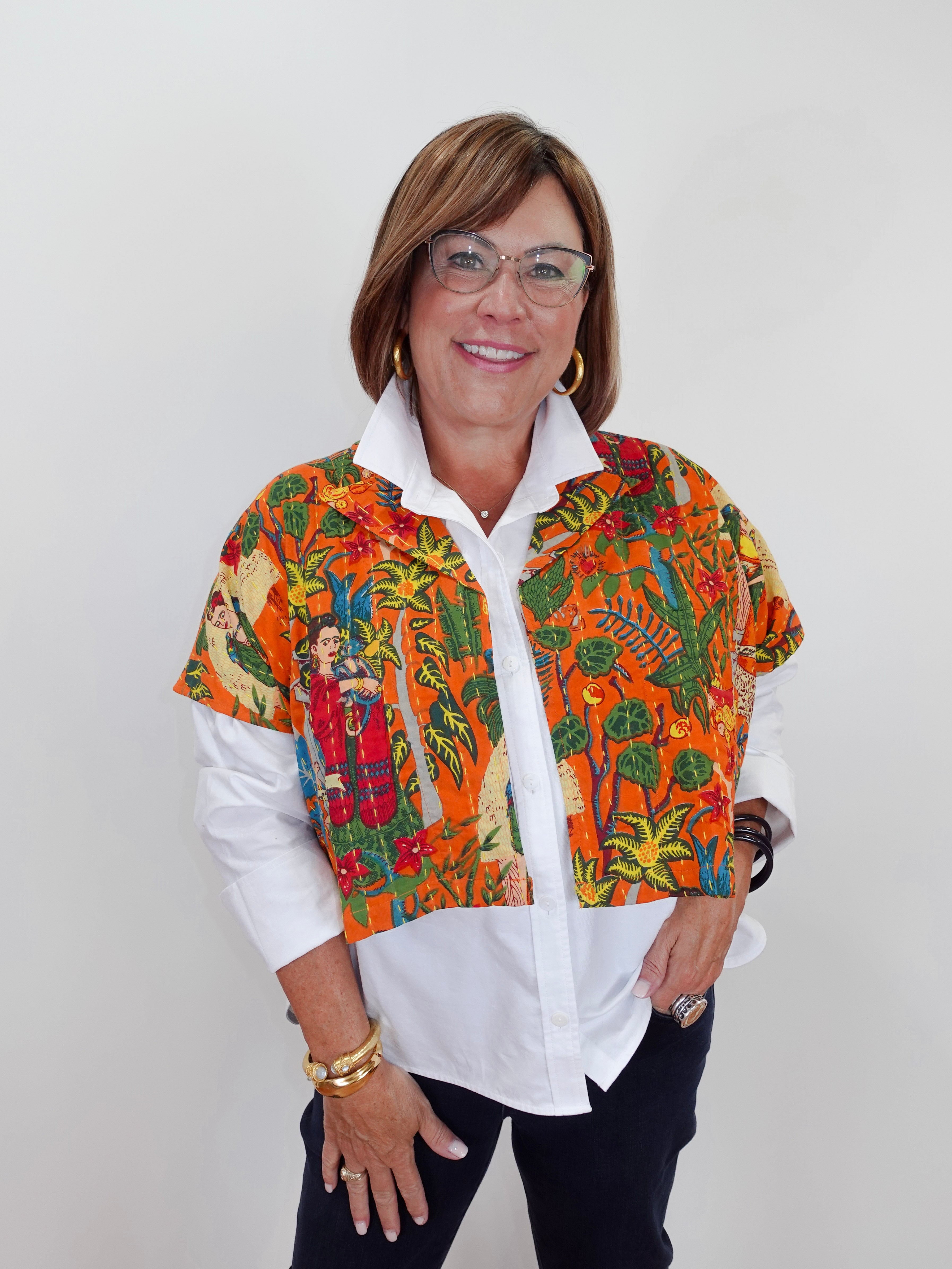 A woman wearing a short-sleeve button-up shirt with a vibrant Mexican Tehuana-style print in orange, green, and red, layered over a white collared shirt and dark pants.
