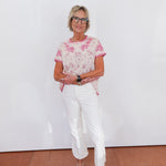 Woman wearing a pink floral top and white pants against a white wall.