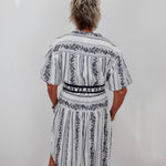 A person viewed from behind wearing a short-sleeve white dress with black vertical stripes and black floral border patterns, standing against a plain white wall.