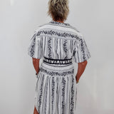 A person viewed from behind wearing a short-sleeve white dress with black vertical stripes and black floral border patterns, standing against a plain white wall.