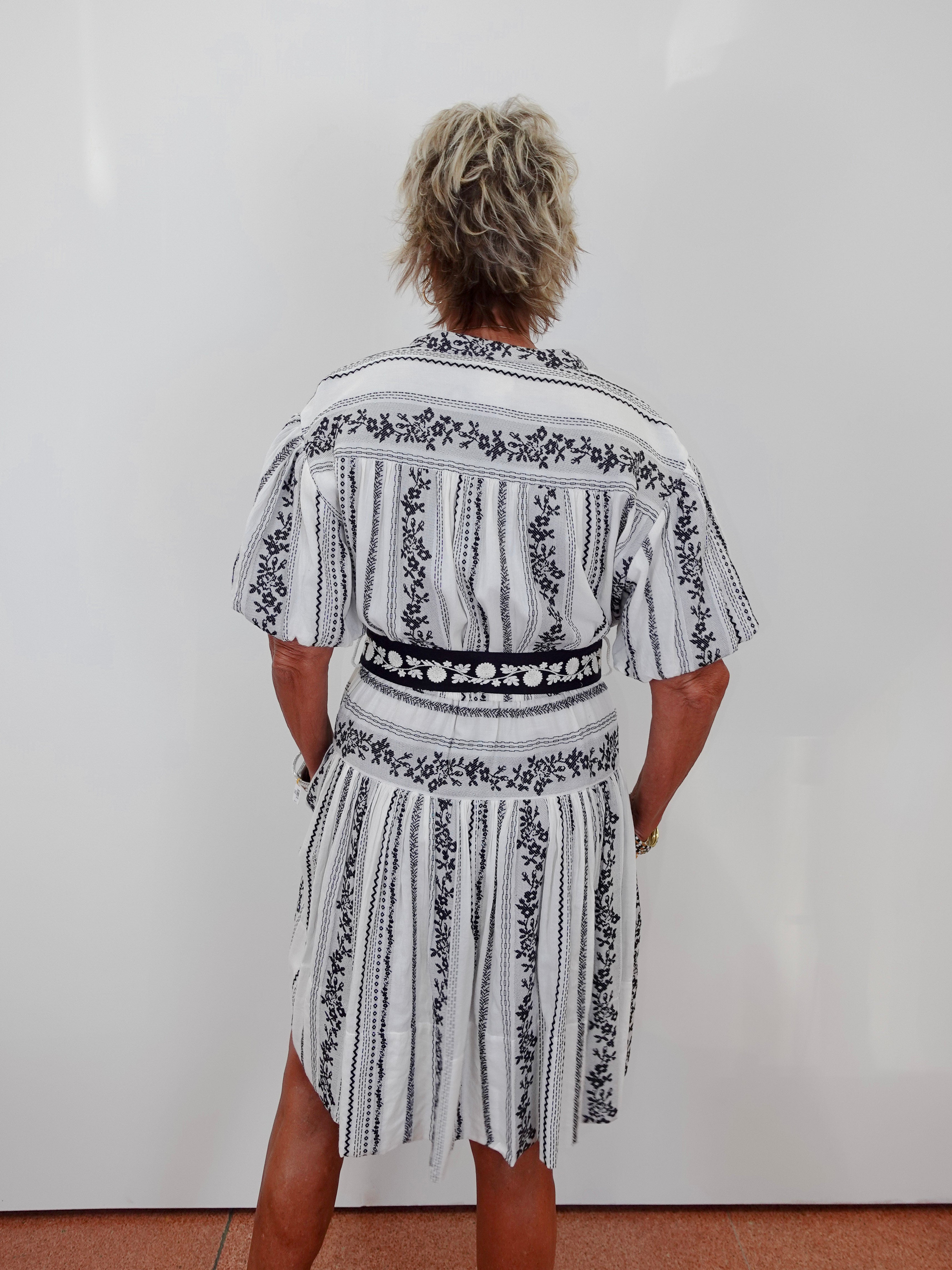 A person viewed from behind wearing a short-sleeve white dress with black vertical stripes and black floral border patterns, standing against a plain white wall.