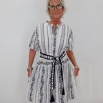 A woman with short blonde hair and glasses wearing a white and black vertically striped dress with a black patterned belt, standing against a plain white wall.