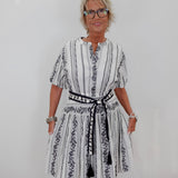 A woman with short blonde hair and glasses wearing a white and black vertically striped dress with a black patterned belt, standing against a plain white wall.