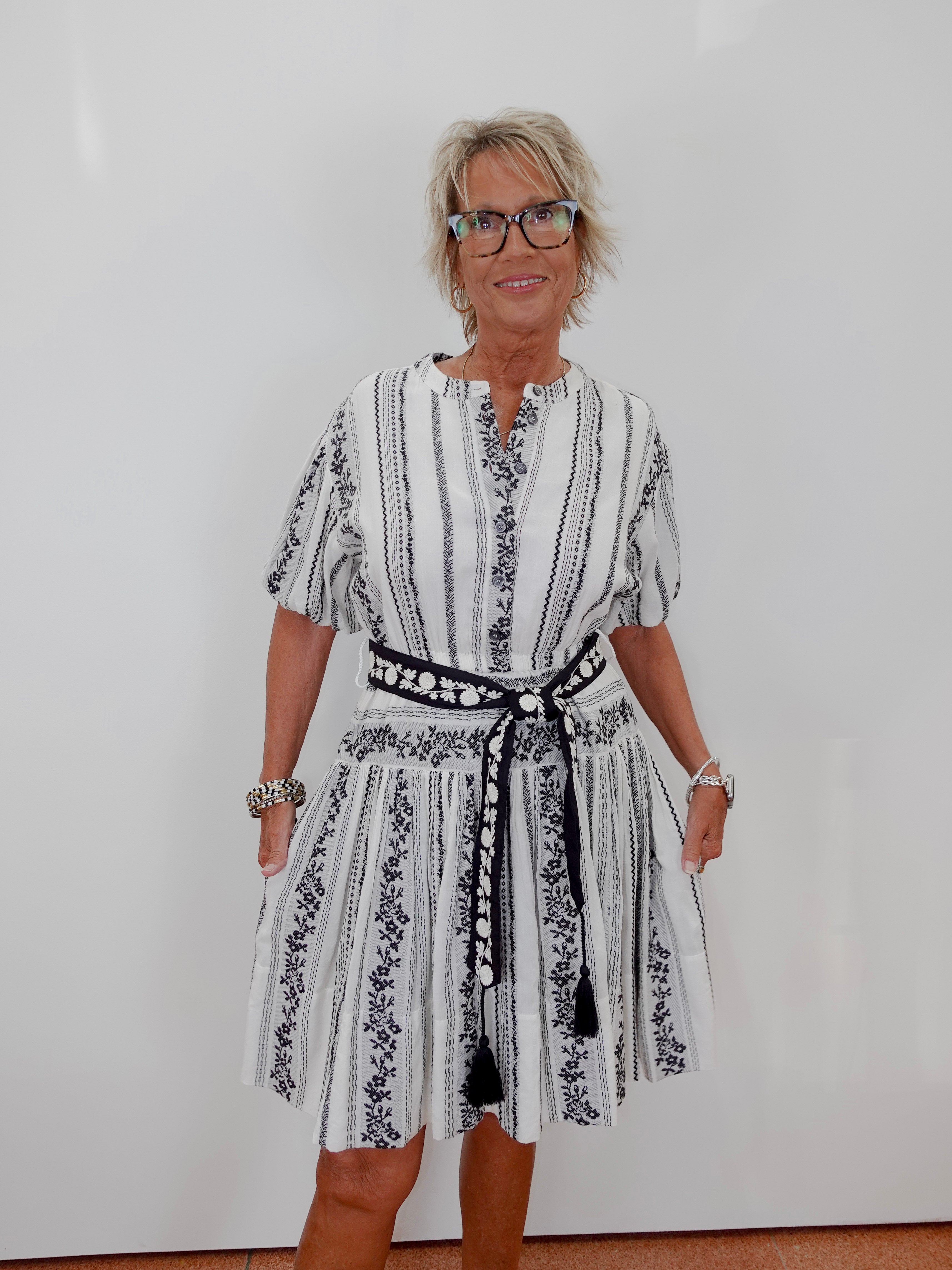 A woman with short blonde hair and glasses wearing a white and black vertically striped dress with a black patterned belt, standing against a plain white wall.