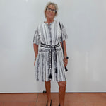 A woman with short gray hair and glasses stands against a white wall wearing a white short-sleeve dress with black vertical stripes and a black patterned belt, paired with black pointed-toe flats.