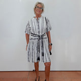 A woman with short gray hair and glasses stands against a white wall wearing a white short-sleeve dress with black vertical stripes and a black patterned belt, paired with black pointed-toe flats.