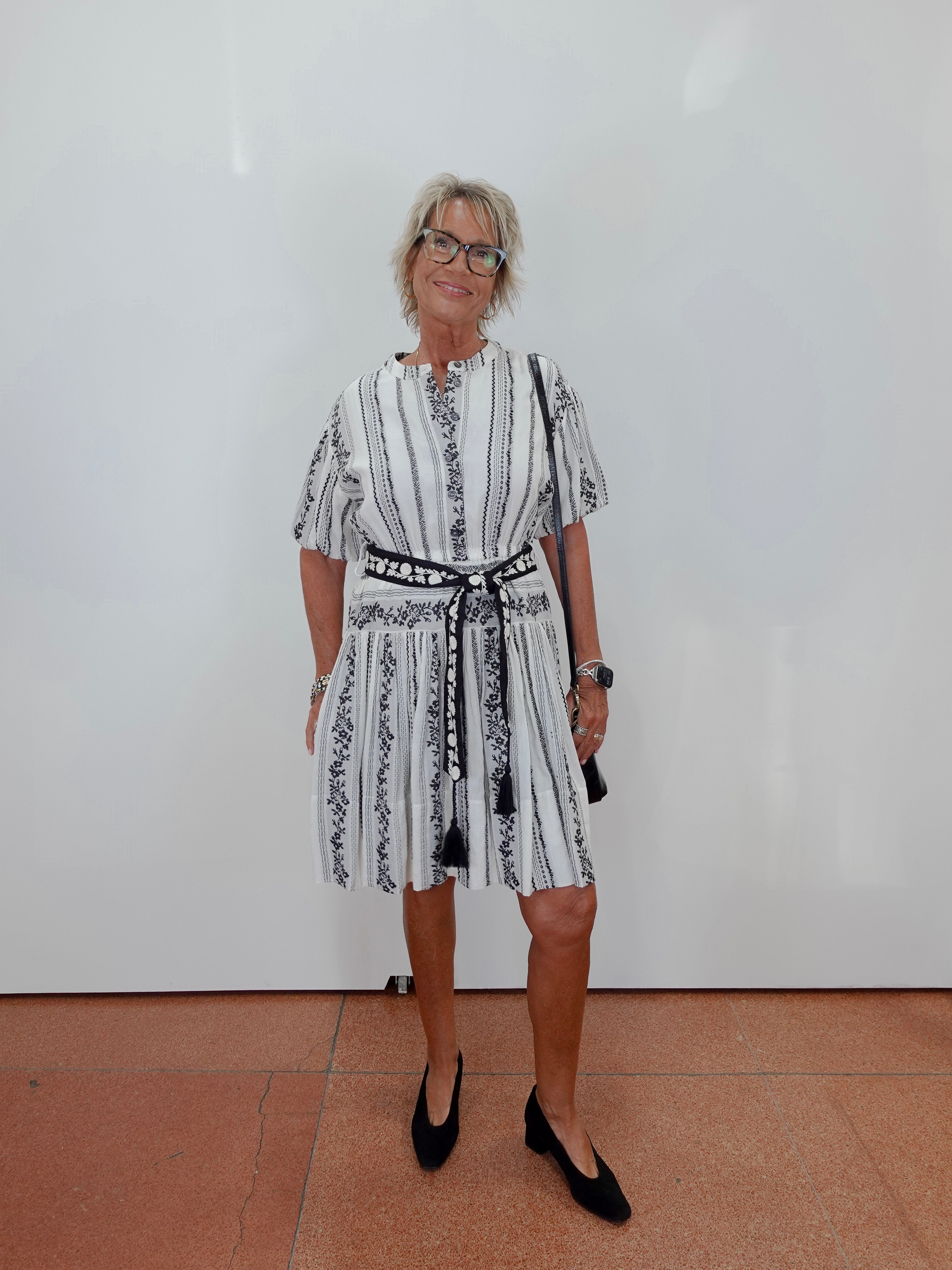 A woman with short gray hair and glasses stands against a white wall wearing a white short-sleeve dress with black vertical stripes and a black patterned belt, paired with black pointed-toe flats.