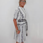 A woman with short blonde hair and glasses wearing a white short-sleeve dress with black vertical stripes and black floral embroidery, standing against a plain white wall.